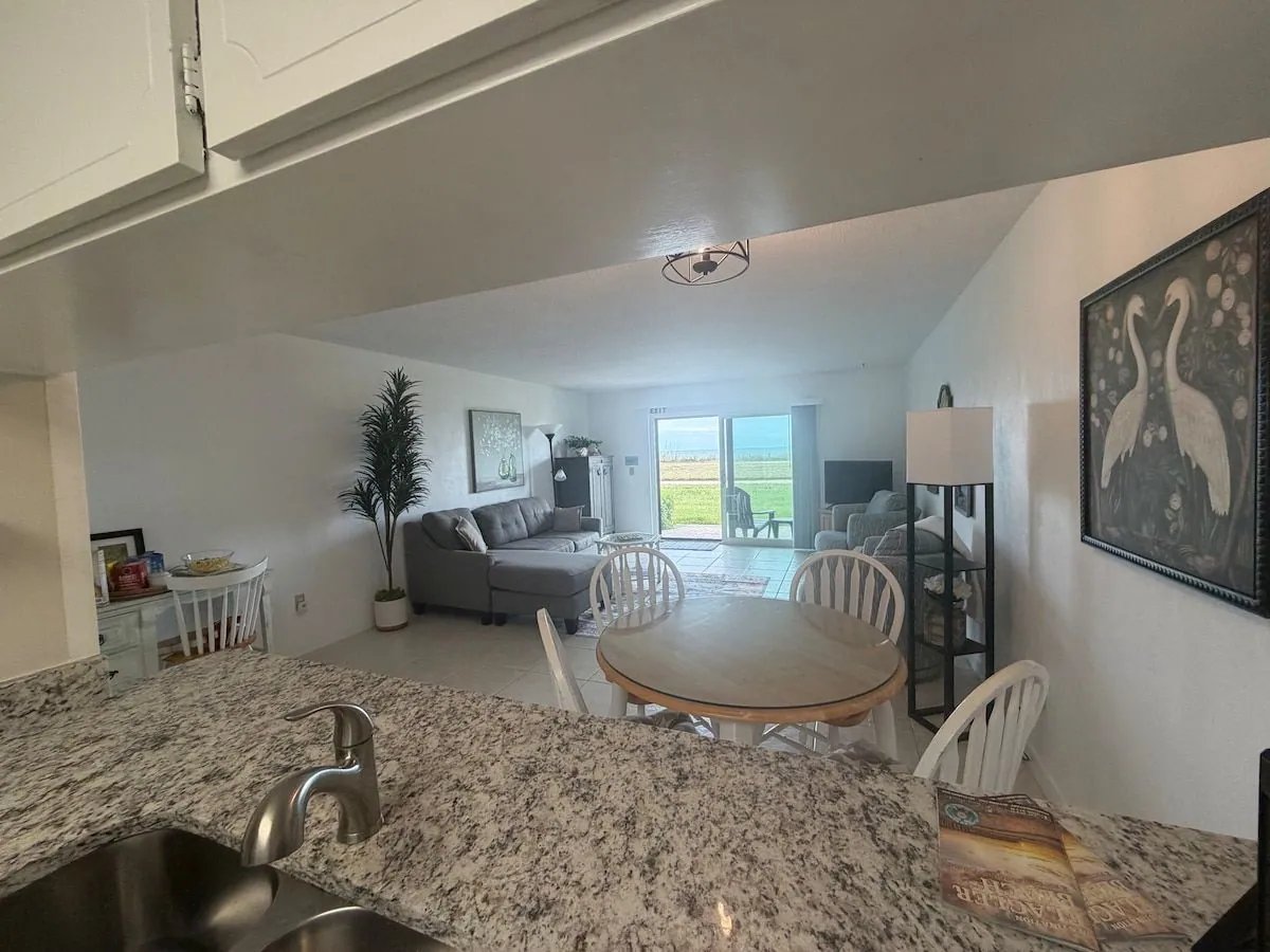 Dining table and living area seen from the kitchen breakfast bar, with the ocean visible through the sliding doors