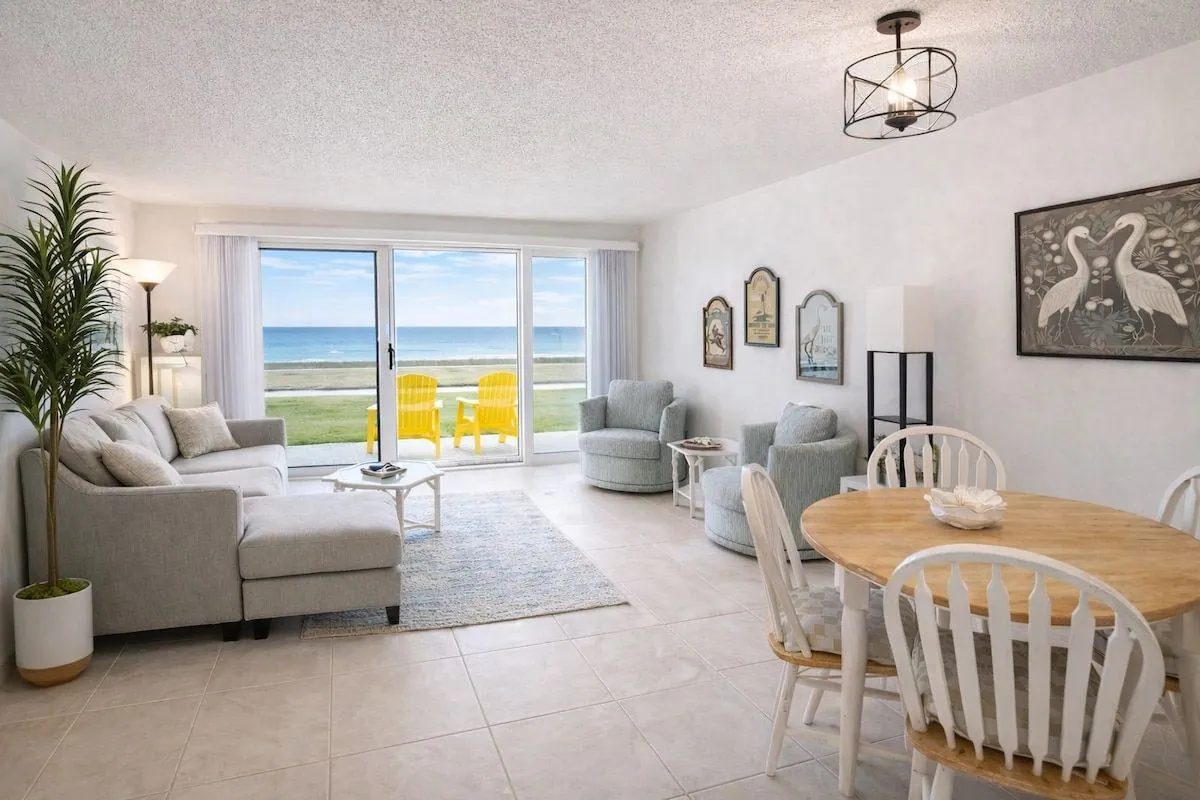 Bright open-plan living and dining area with sliding glass doors framing a full Atlantic Ocean view and yellow chairs out on the lawn