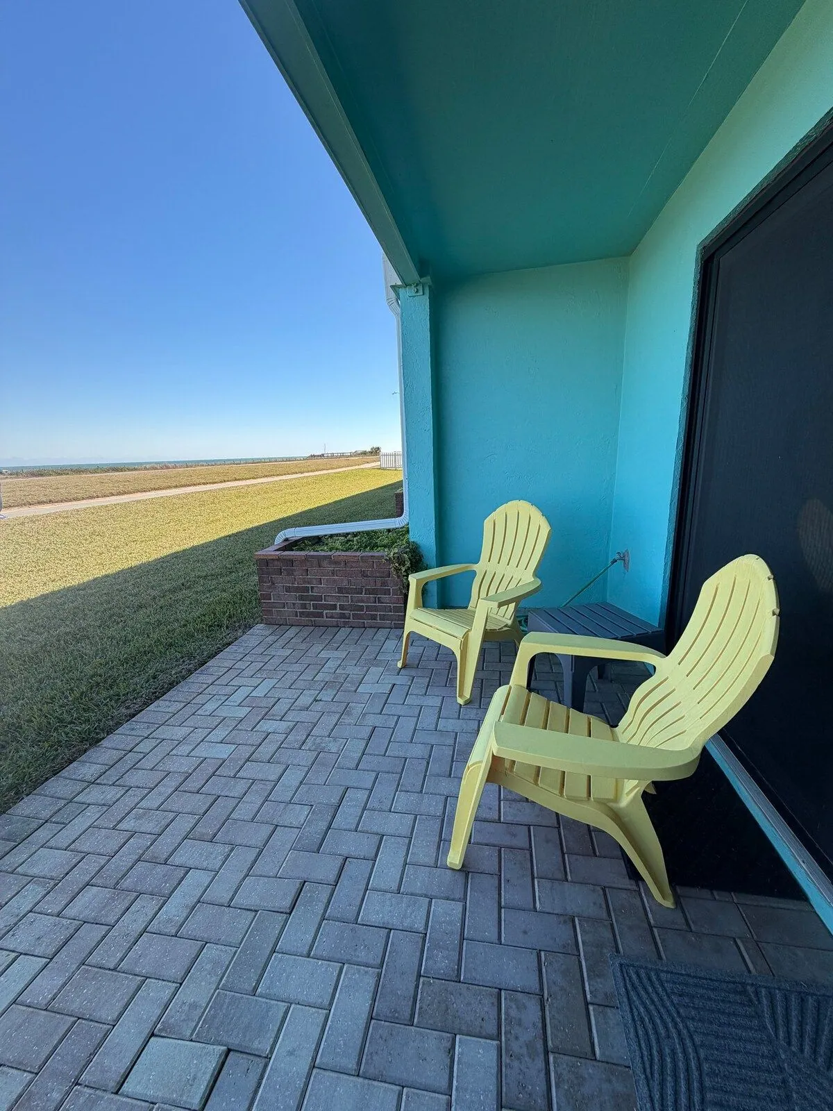 Covered lower patio with yellow Adirondack chairs looking across the lawn and beachfront walking path to the ocean
